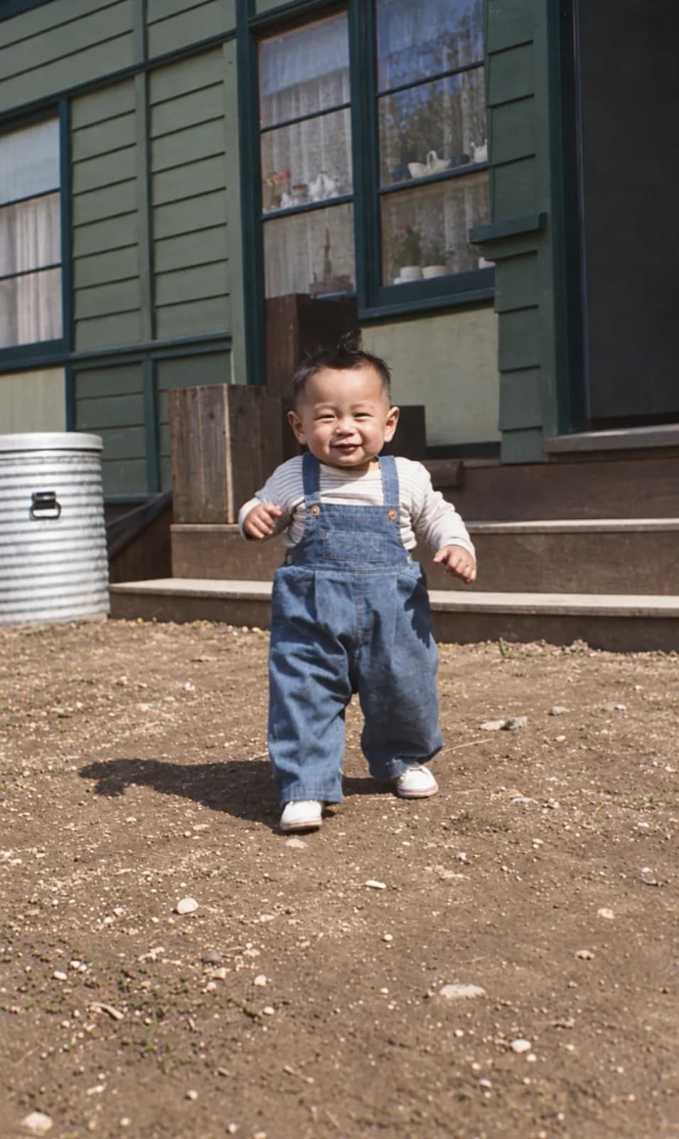 Restored: toddler in denim overalls walking toward the camera in front of a green house