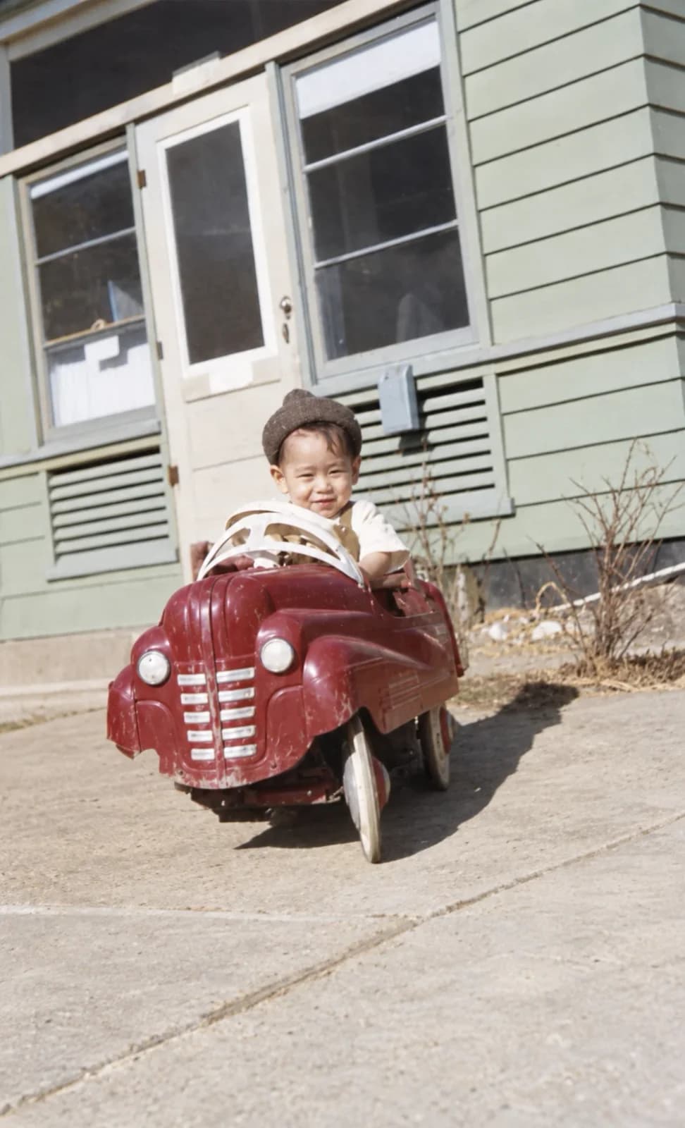 Restored: toddler in a cap sitting in a red pedal car in front of a house