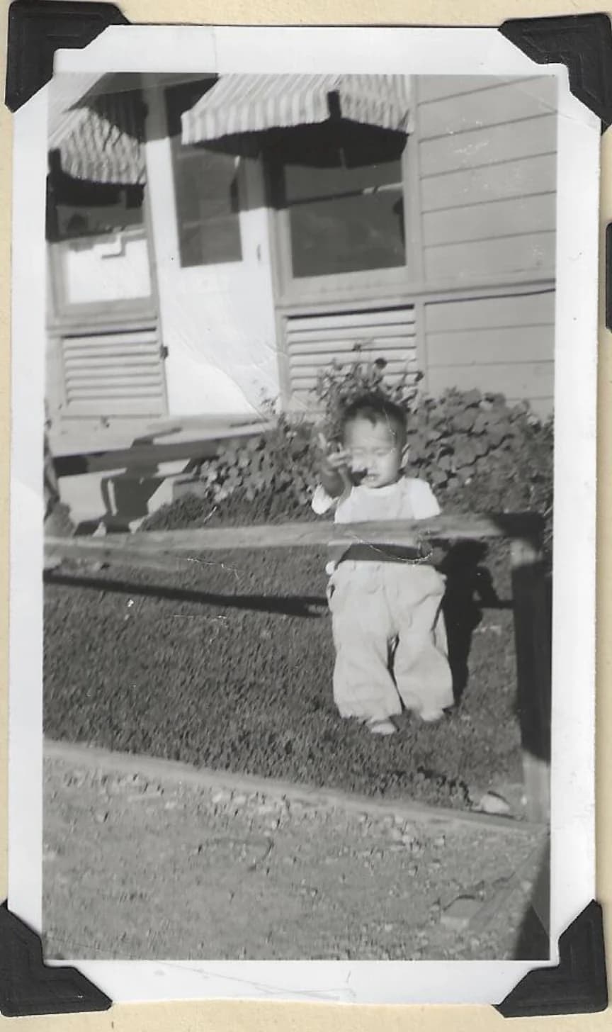 Original: black-and-white snapshot of a toddler behind a wooden fence, in black photo corners